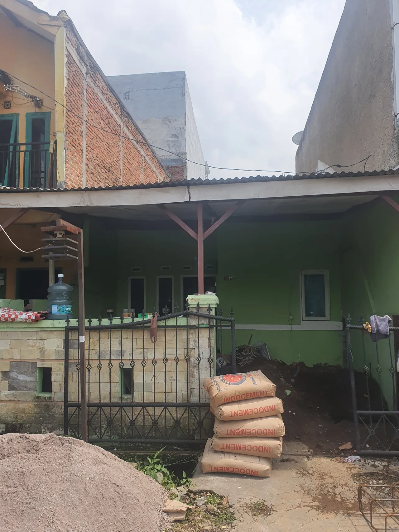 Rumah satu lantai berdinding hijau sebelum renovasi, dengan material bangunan menumpuk di halaman